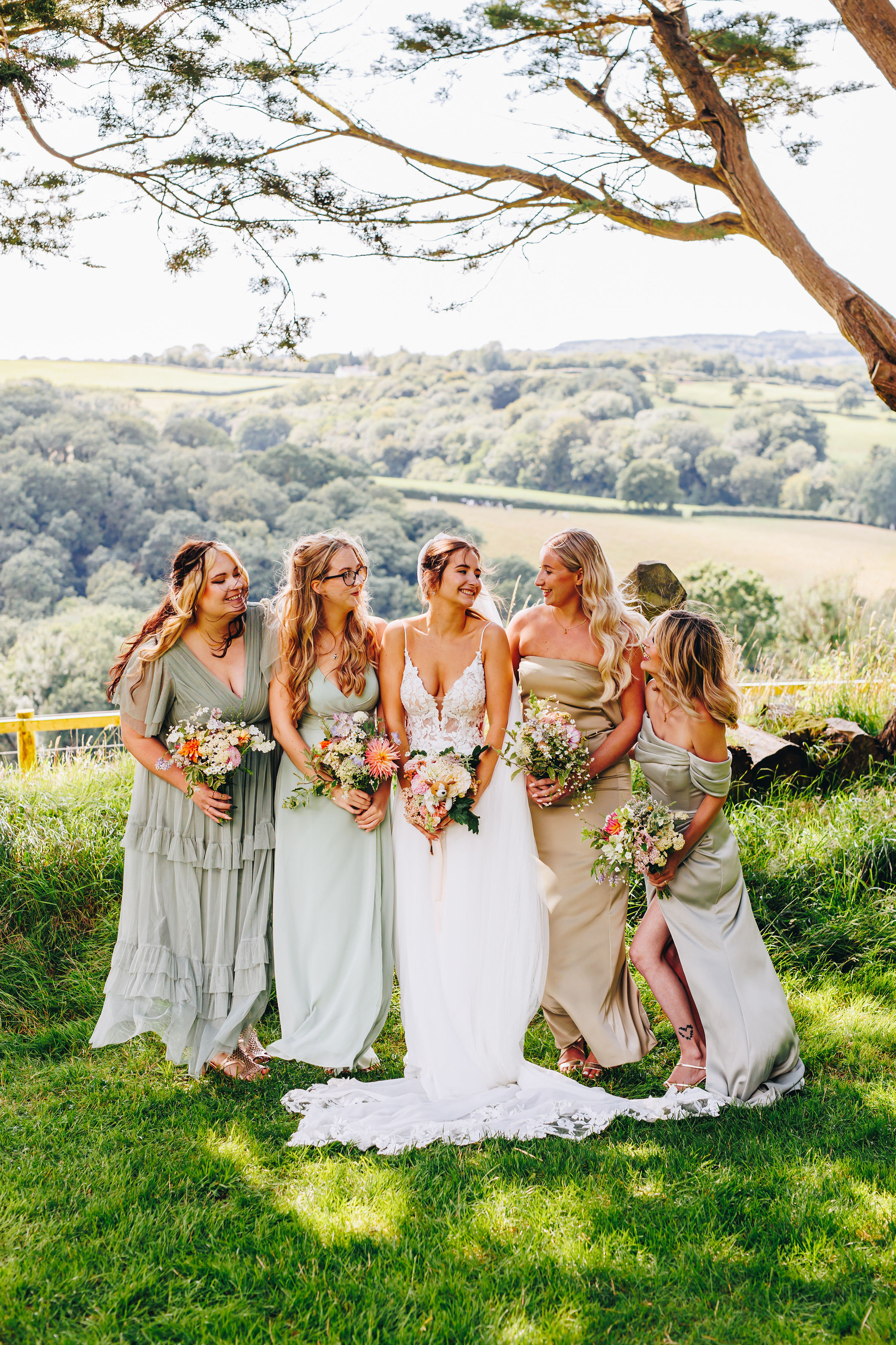Bride and bridesmaids smile at each other, stood under trees