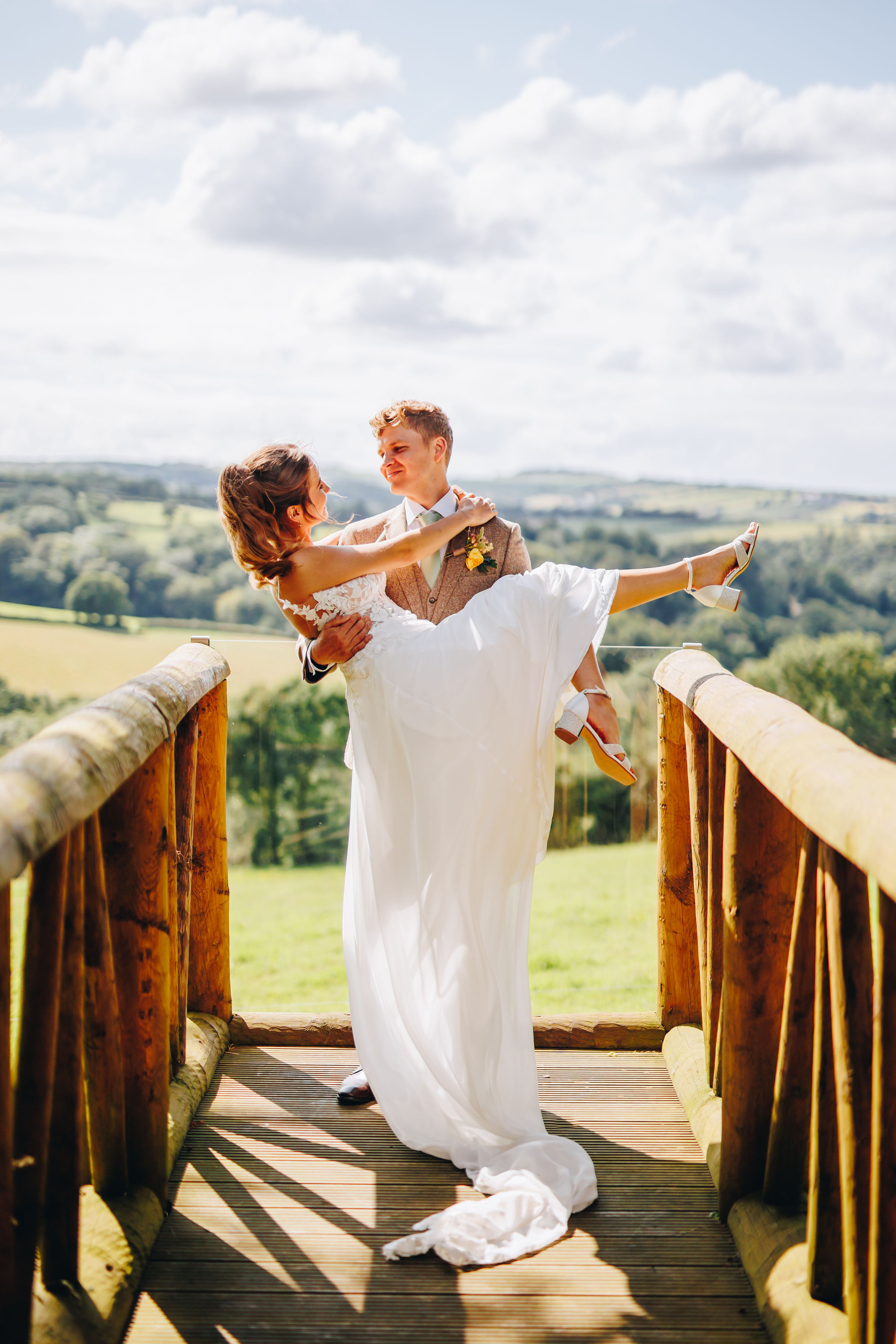 Bride being held up by groom with dress trailing on the floor, on a wooden path with a valley behind them
