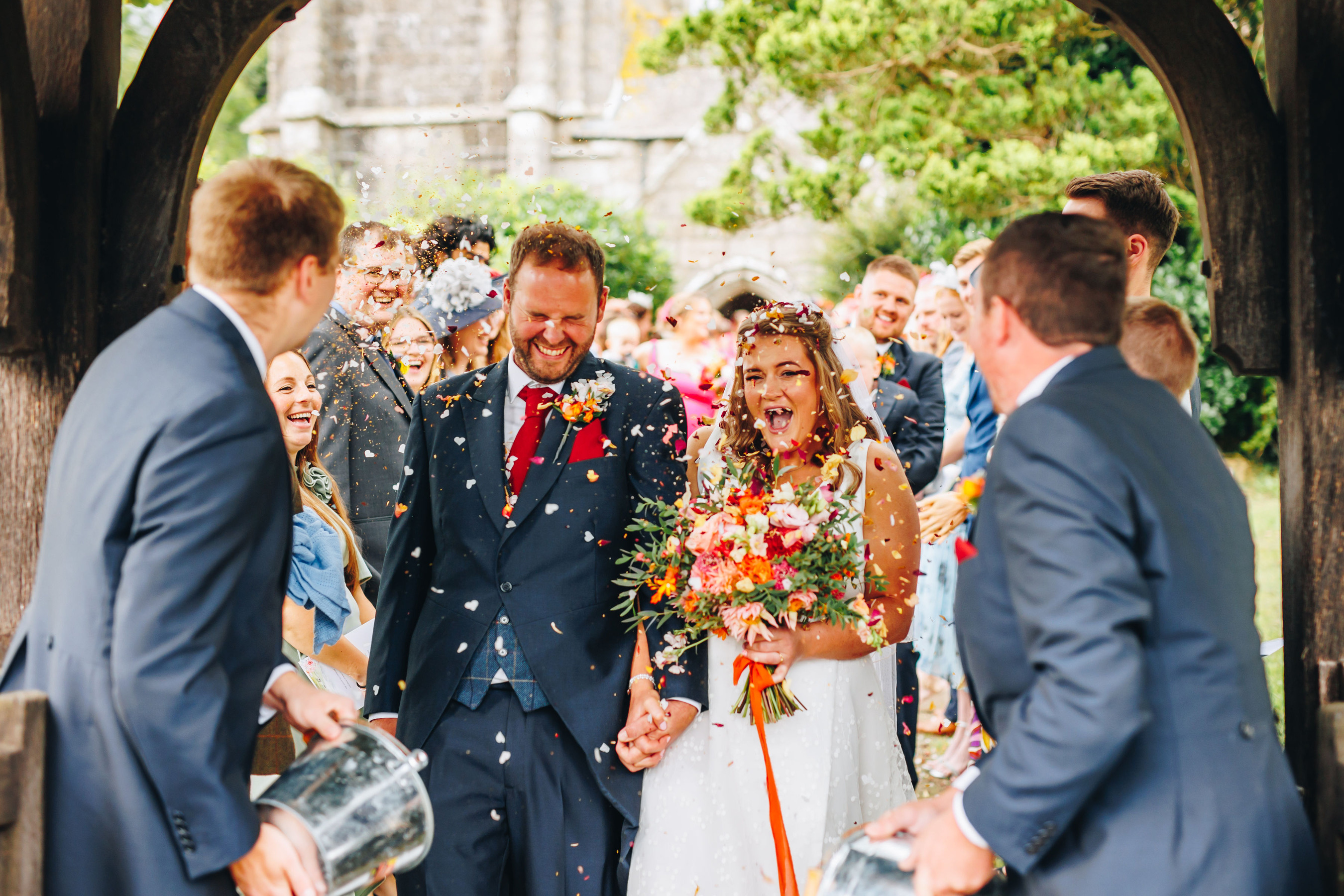 Bride and groom stood in a shower of confetti, with two groomsmen in the foreground holding buckets of it
