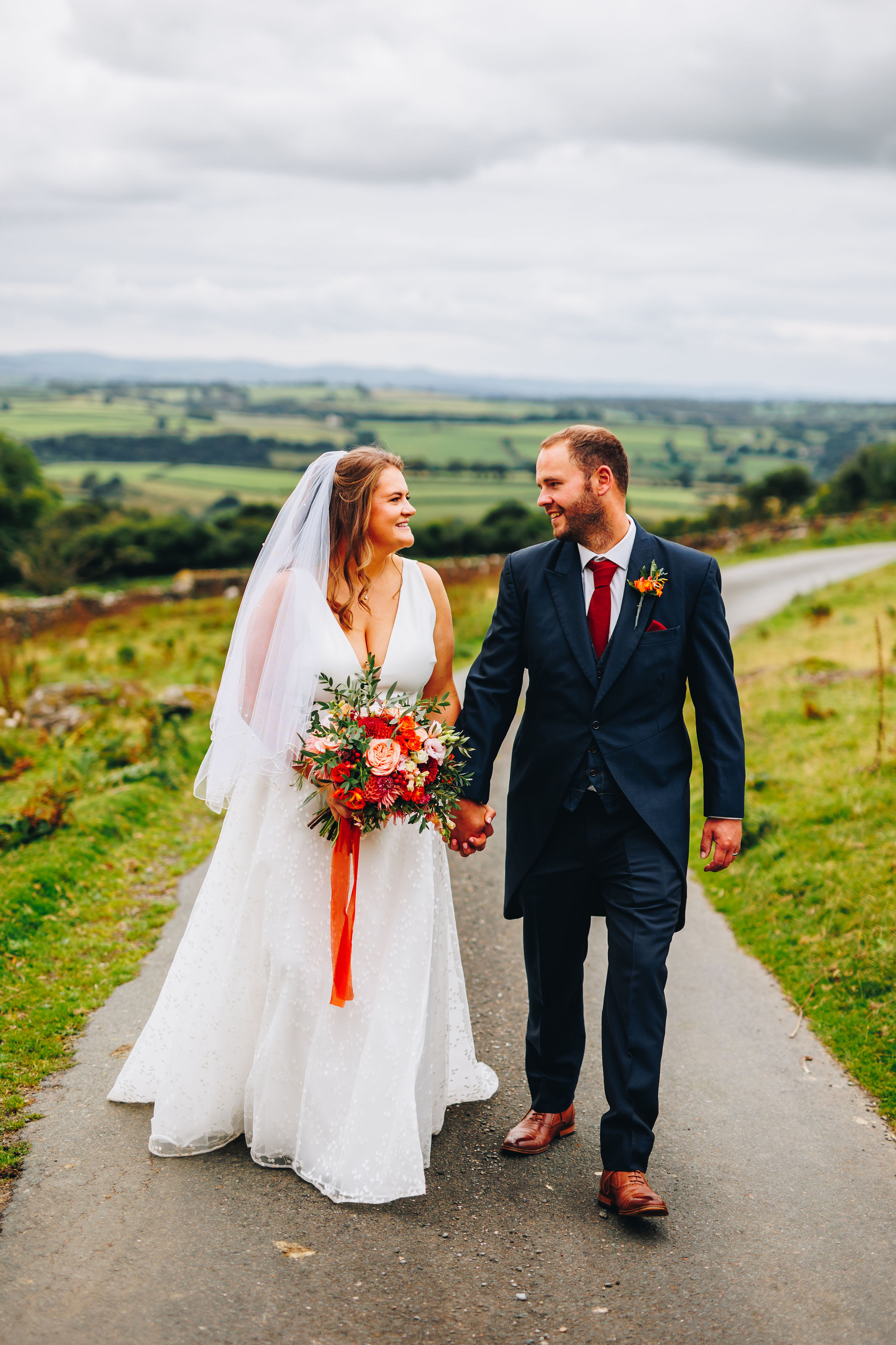 Bride and groom walk up a farm track, holding bouquet and smiling at each other