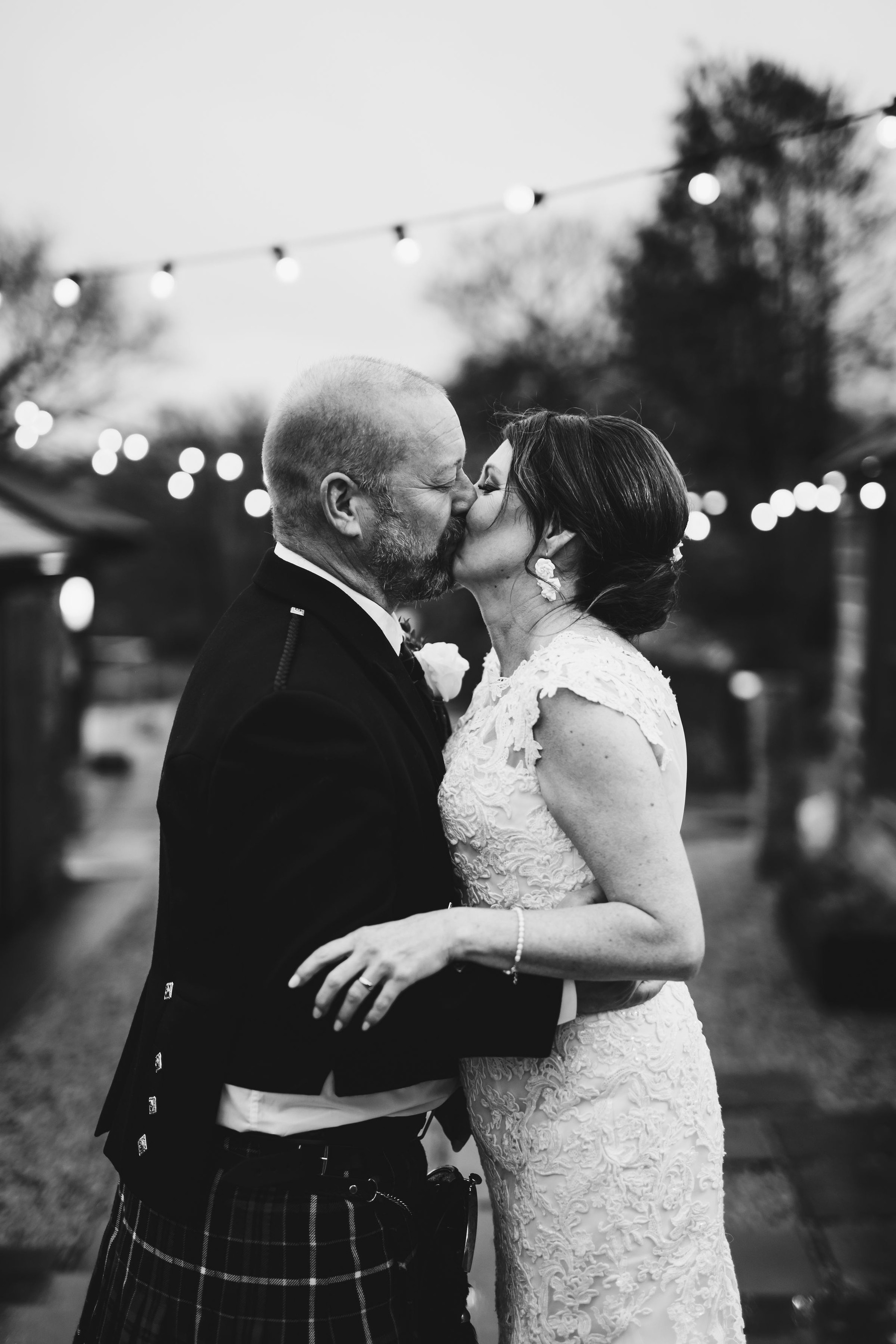 Black and white photo of couple kissing with fairy lights in the background