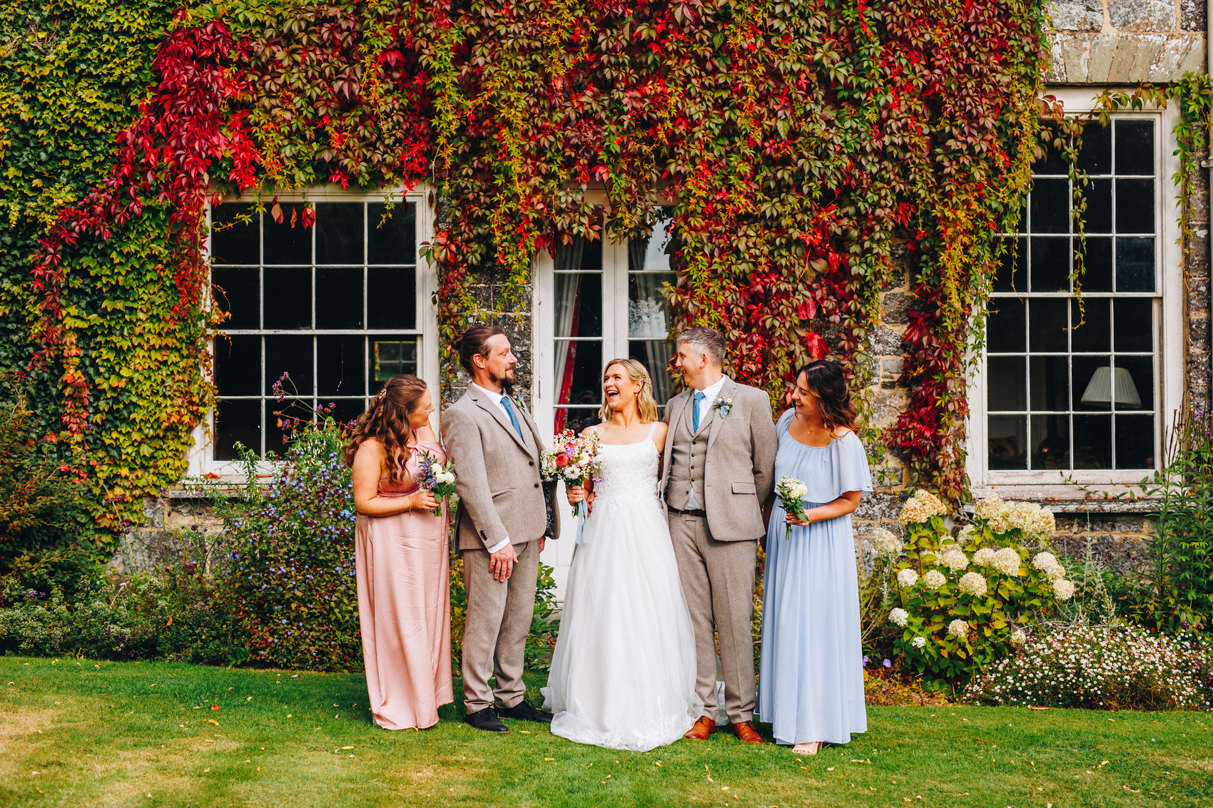 Group photo of couple and bridal party in front of an ivy-covered building, smiling