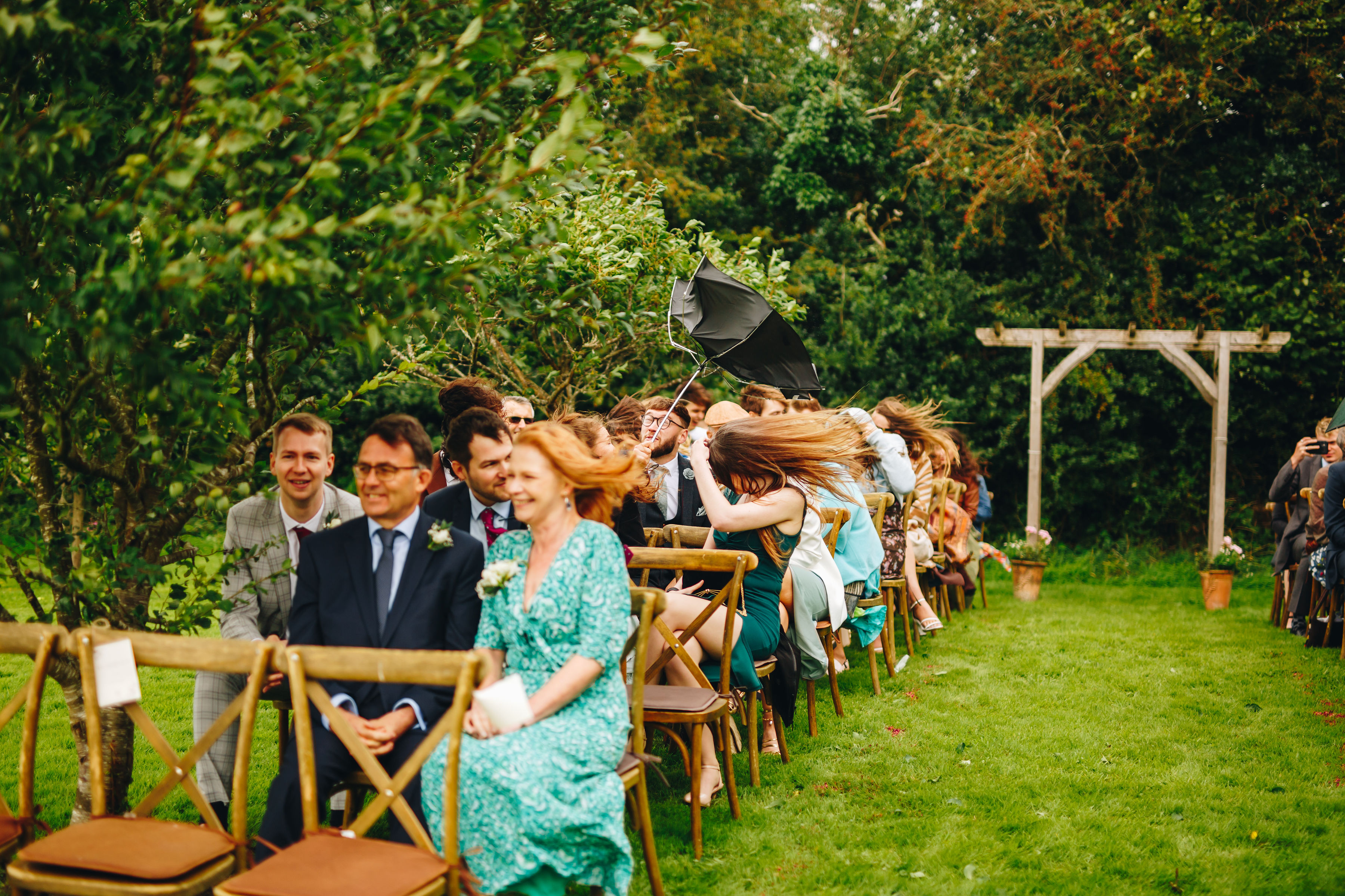 Guests sat at a outdoor wedding ceremony, wind making hair go horizontal and umbrellas go inside out