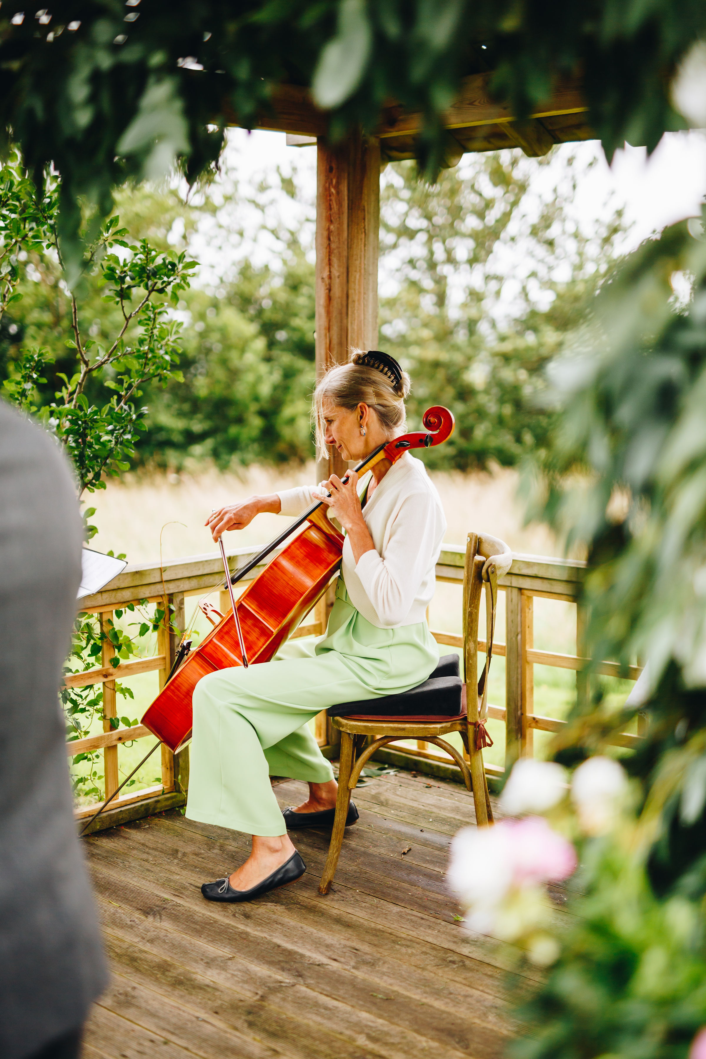 Cellist playing at an outdoor ceremony in the breeze, surrounded by greenery