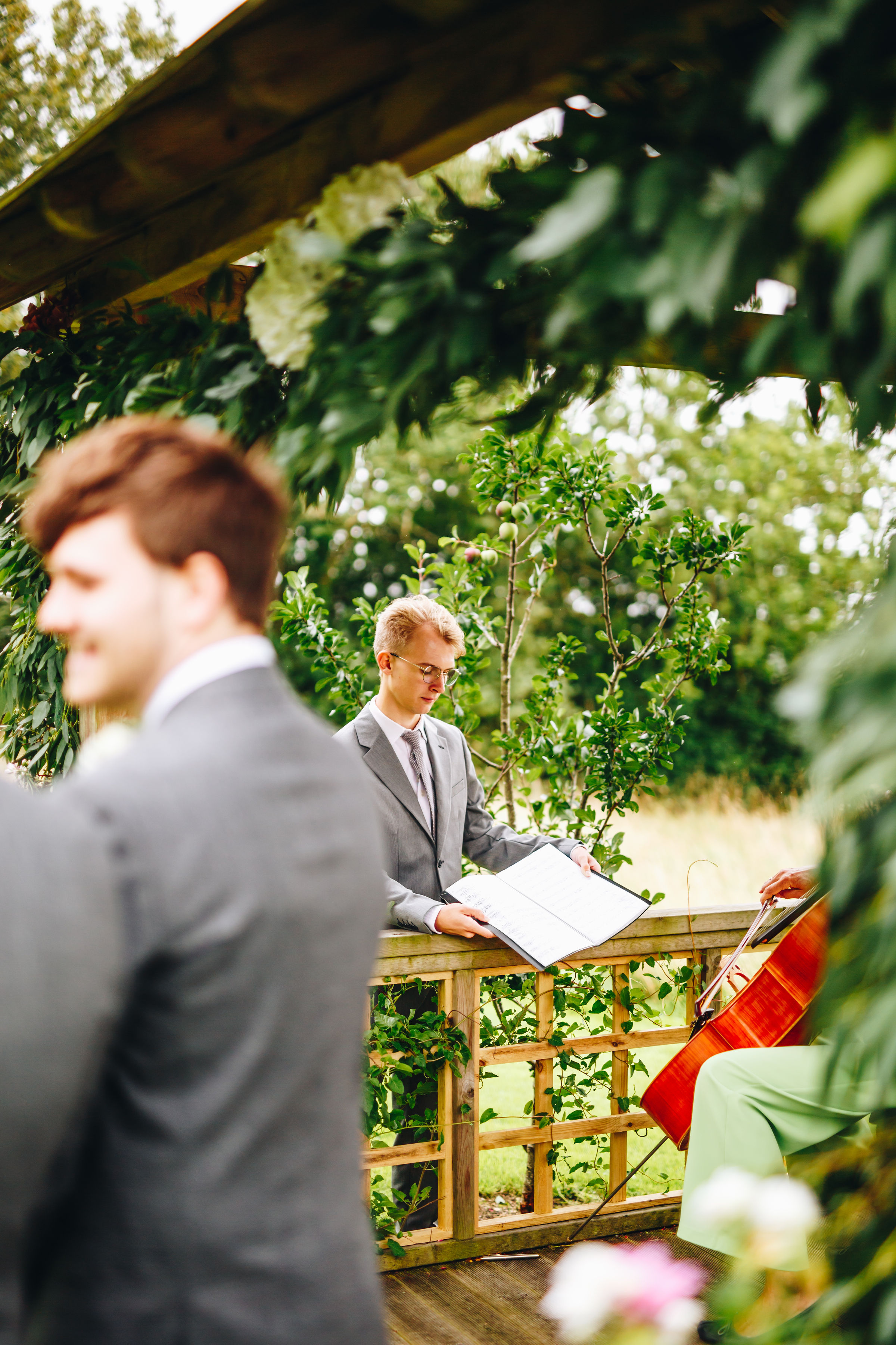 Groomsman holds out the cellist's music book, surrounded by greenery