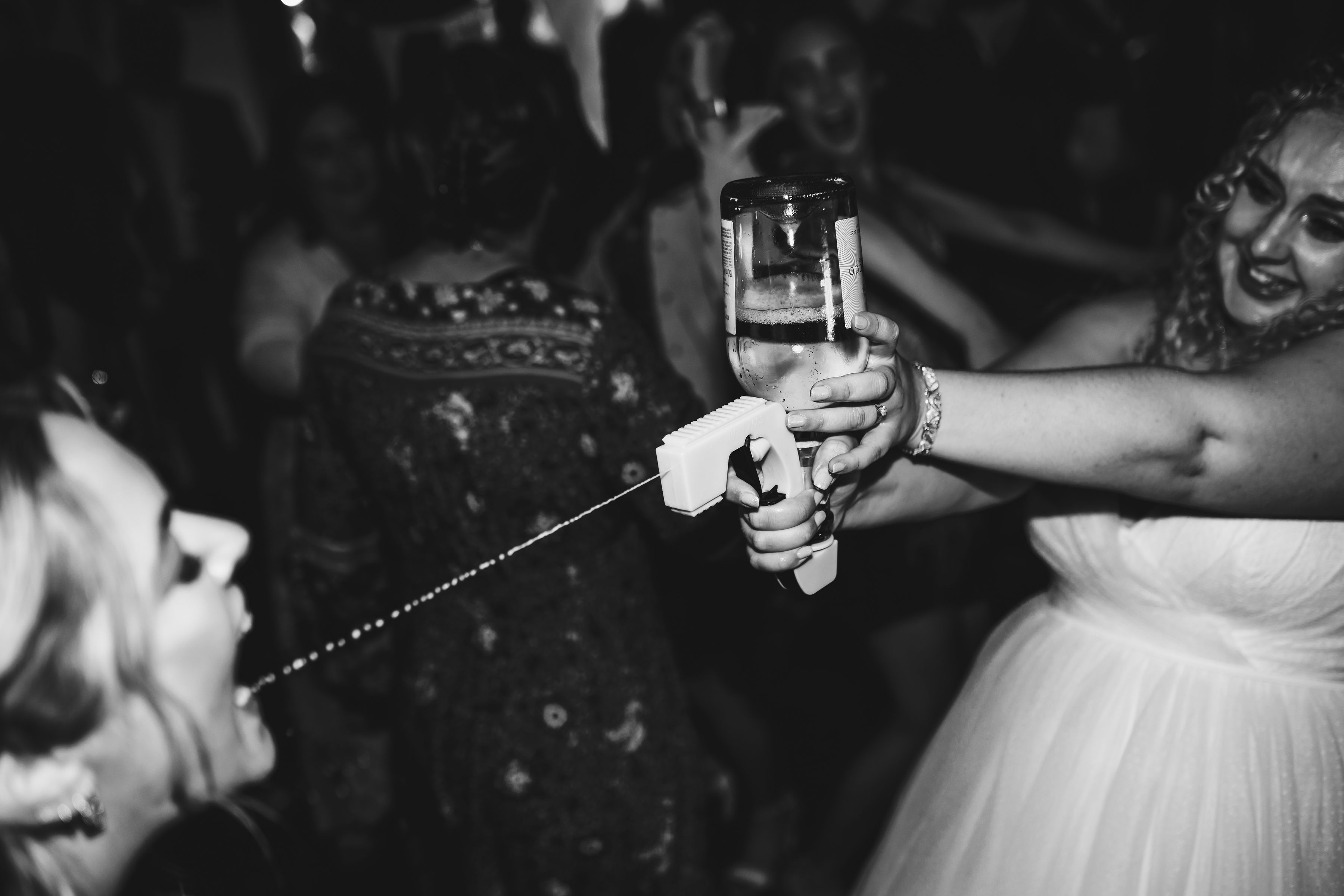 Black and white photo of bride using Prosecco gun to spray drink into guest's mouth