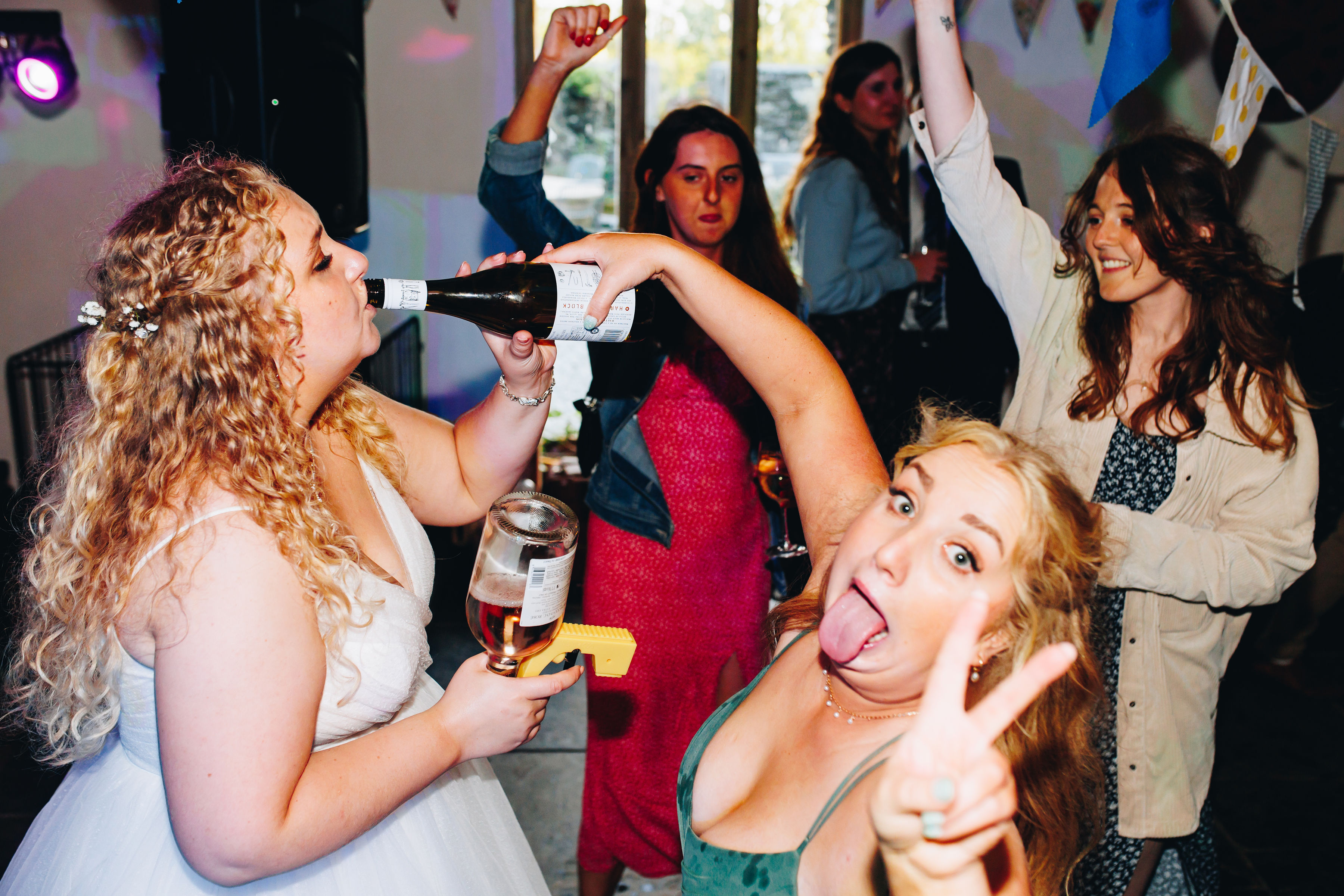 Bridesmaid holding bottle to bride's mouth while guests party behind them