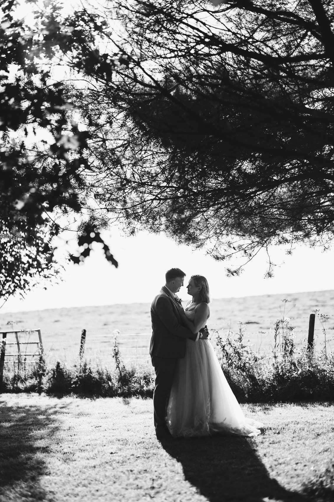 Black and white photo of bride and groom snuggling together under the trees near sunset