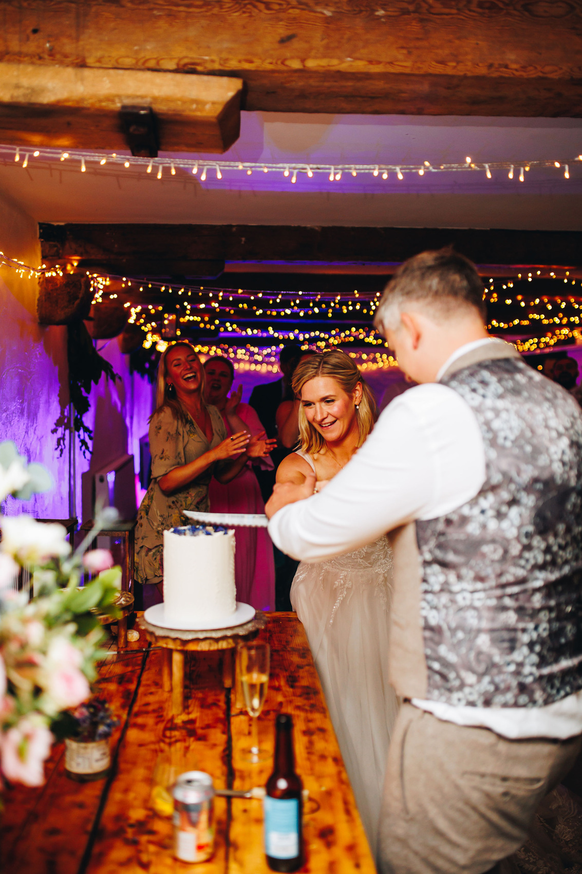 A bride and groom cutting their white wedding cake in a room with wooden beams and fairy lights
