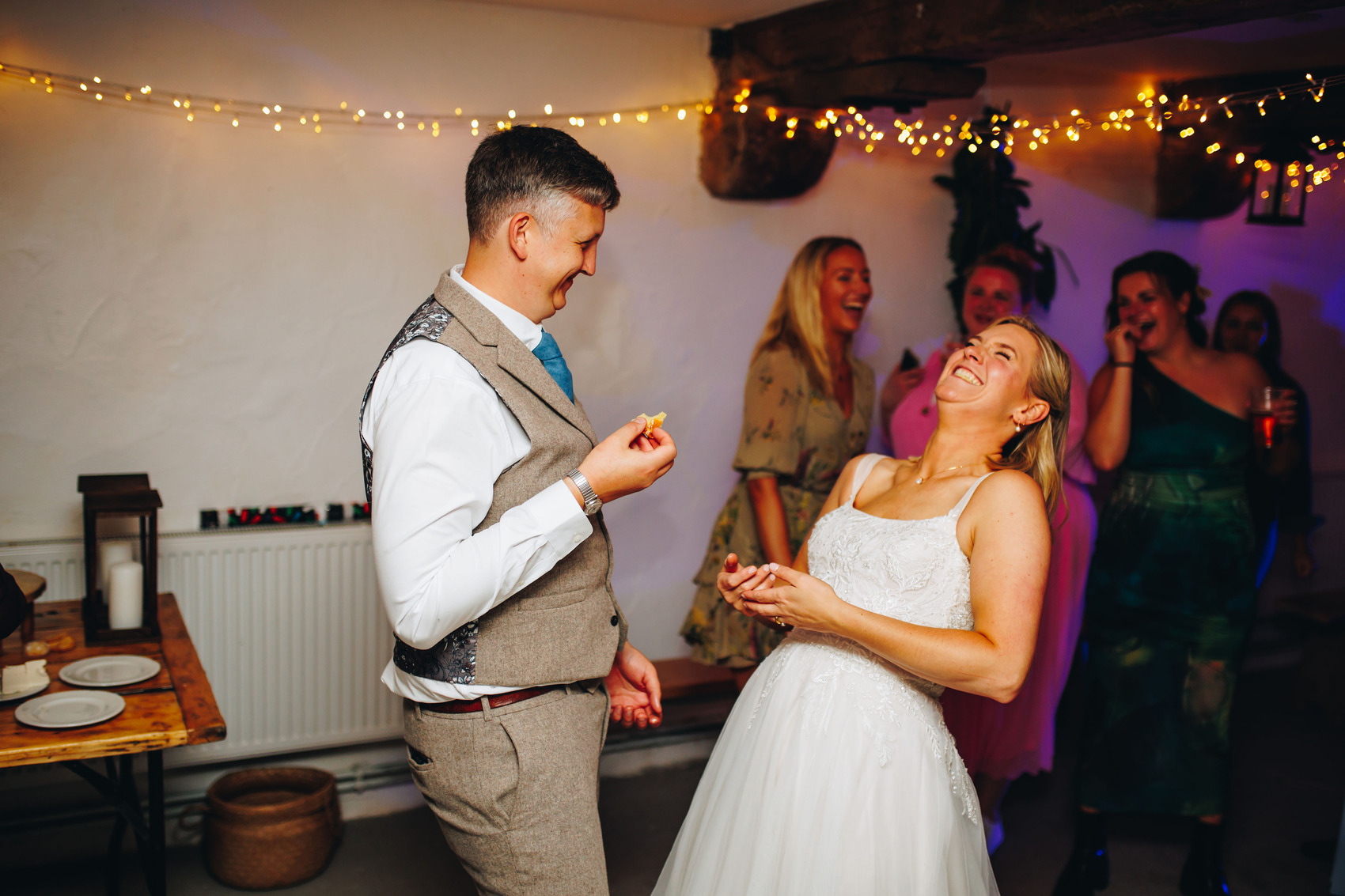 Bride and groom laughing while eating their wedding cake surrounded by smiling friends