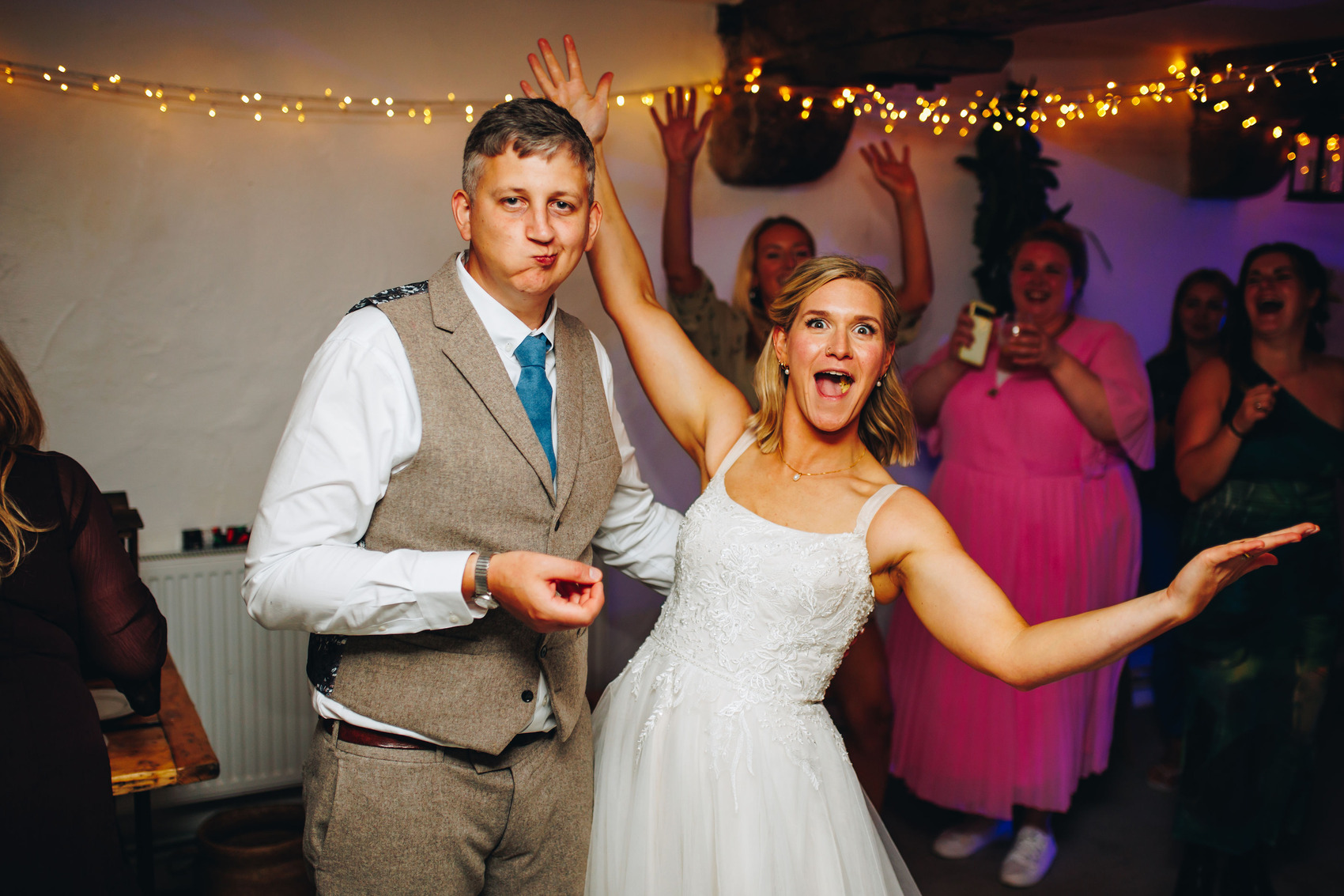 Bride looking amazed and groom looking content while eating their wedding cake as guests cheer behind them