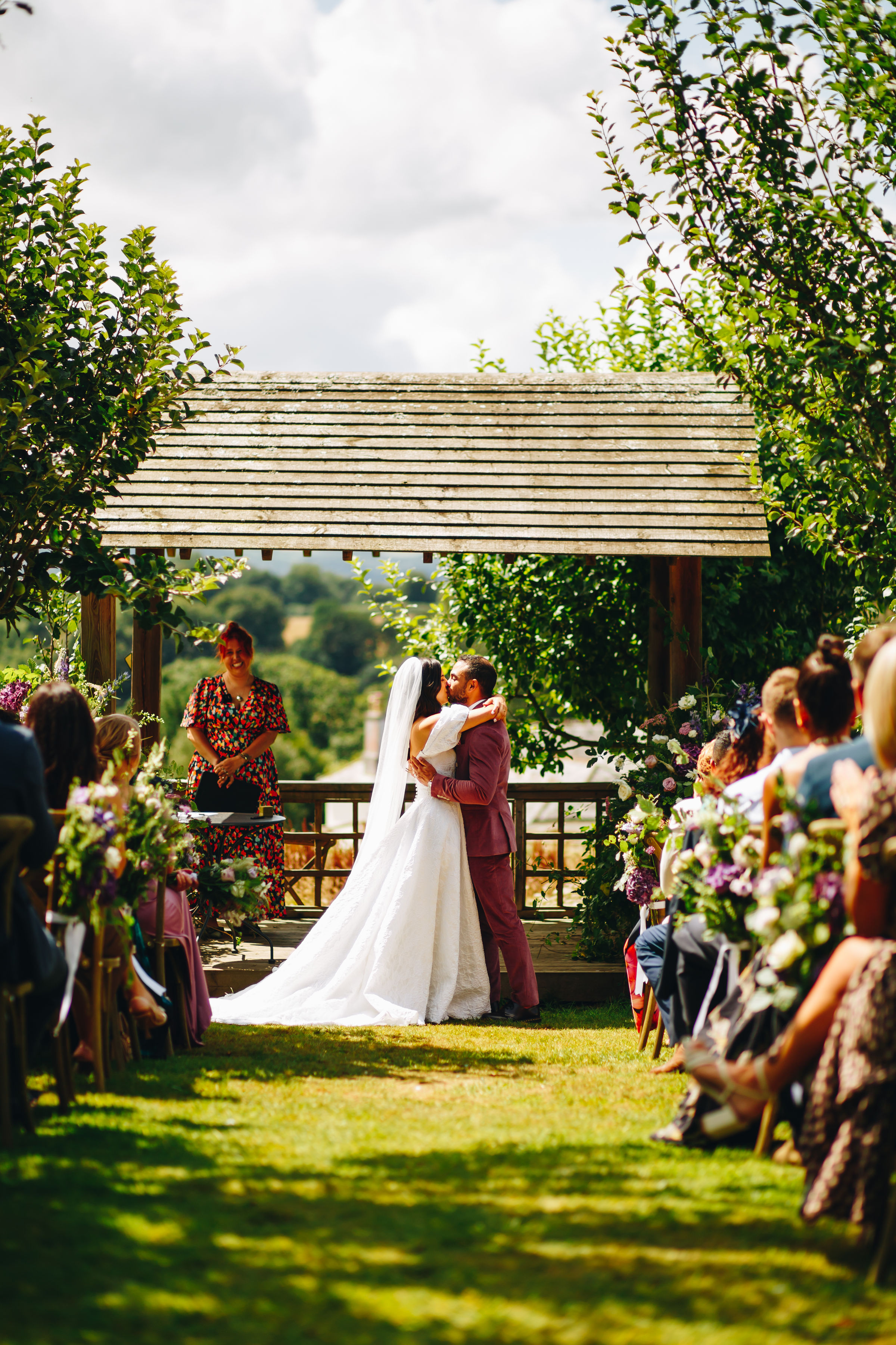 A couple embracing at the end of their wedding ceremony, in front of a wooden structure surrounded by trees